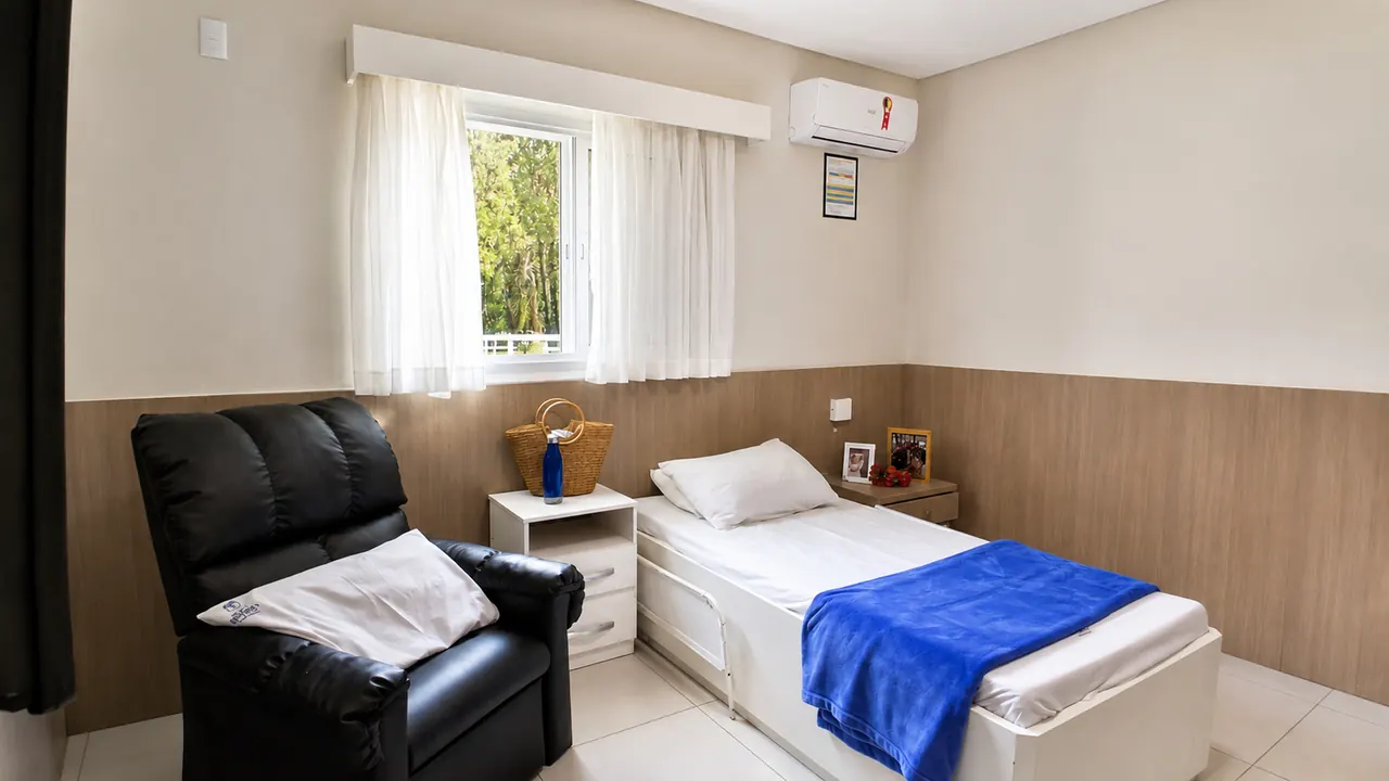 Quarto da Nova Belluno com cama, poltrona e janela para a área verde
